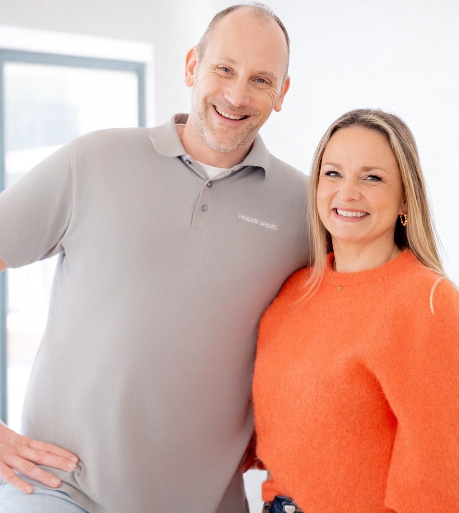 Ein Mann in einem grauen Polo und eine Frau in einem orangefarbenen Pullover lächeln freundlich.